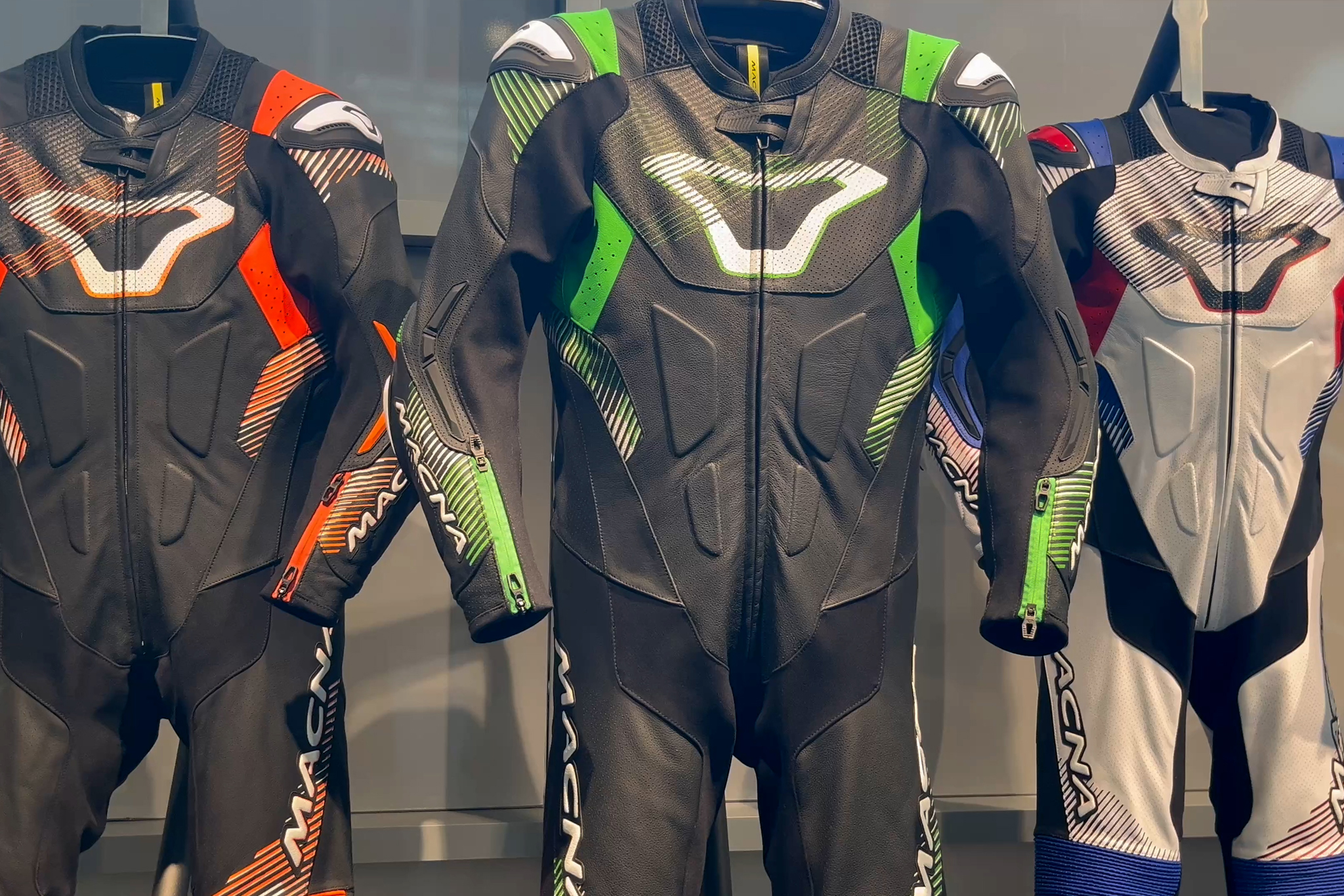Drie kleurrijke leren motorpakken van het merk Macna hangen in een showroom, geschikt voor sportieve rijders.